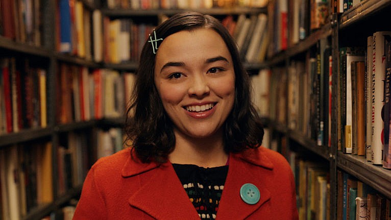 a smiling actor wears a red jacket. there are book shelves behind her. 